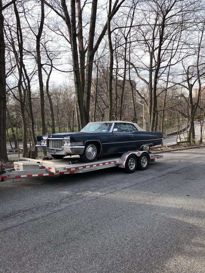 who-we-are Classic blue car on a trailer amidst leafless trees on a street.